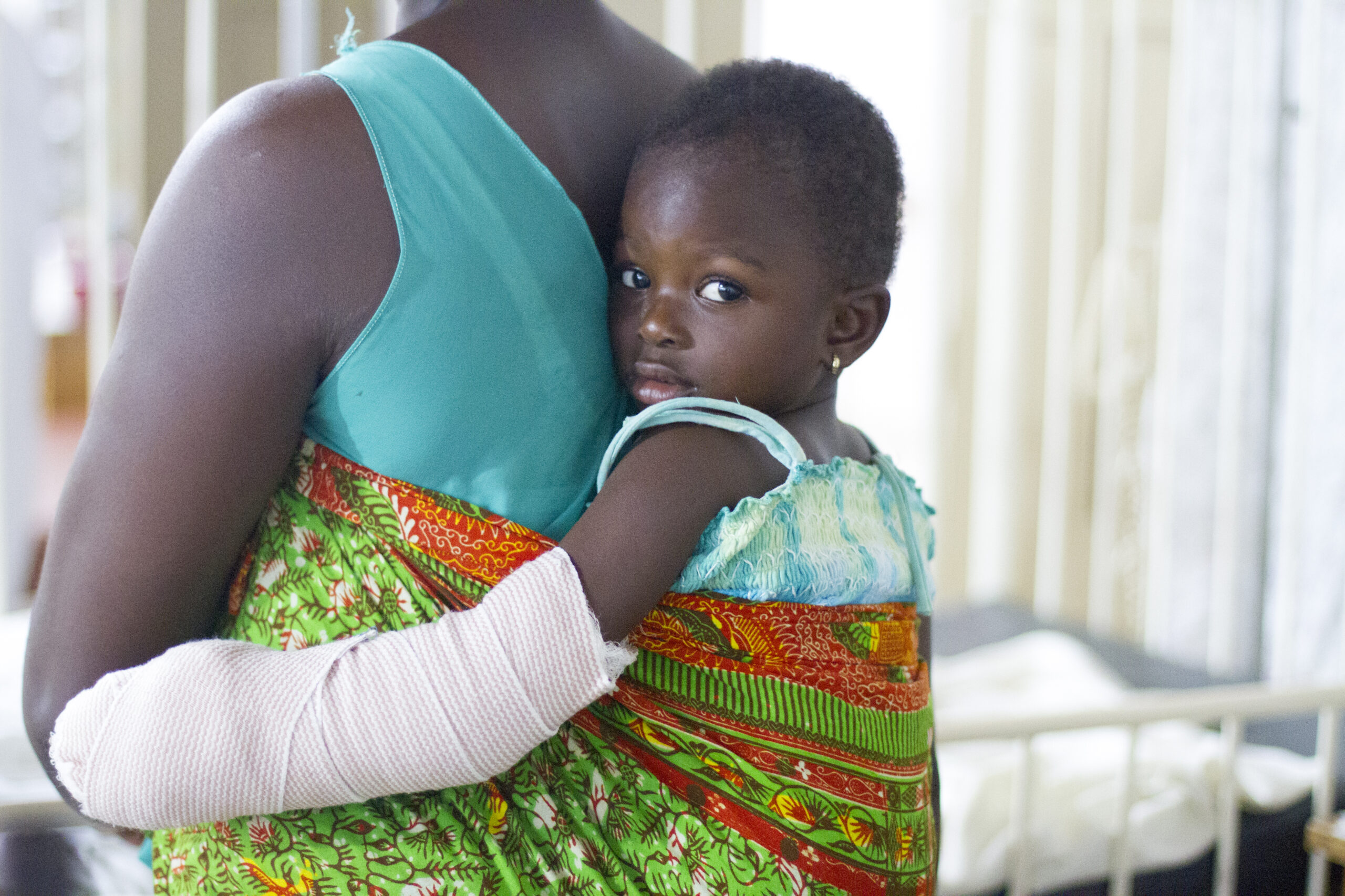 A young patient looks over her mother's shoulder, arm freshly bandaged